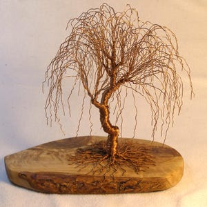 May include: A sculpture of a weeping willow tree crafted from thin copper-colored wire. The tree features a twisted trunk and numerous drooping branches, mounted onto a polished, light-brown wooden base with a natural, irregular edge.