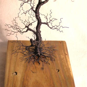 Pode incluir: Uma escultura de arame de uma &aacute;rvore com uma base de madeira marrom. A &aacute;rvore tem um tronco grosso e muitos galhos que s&atilde;o torcidos e encaracolados. As ra&iacute;zes da &aacute;rvore s&atilde;o vis&iacute;veis e se espalham pela base.