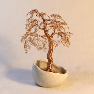 May include: A handcrafted copper wire bonsai tree sculpture in a small, off-white ceramic pot. The tree features a detailed trunk and branches with delicate, looped leaves, creating a miniature, artistic representation of nature.