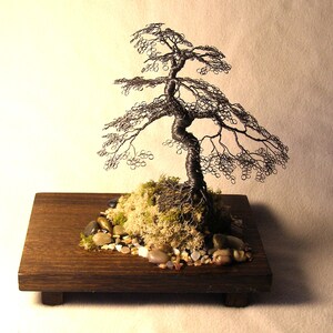 May include: A wire sculpture of a bonsai tree with a brown wooden base and small stones. The tree is made of silver wire and has a realistic shape.