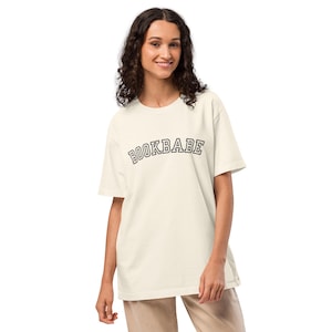 Può includere: T-shirt color crema con la scritta "BOOKBABE" arcuata sul petto in un carattere nero in stile collegiale. La maglietta ha maniche corte e una vestibilità rilassata. La modella indossa la maglietta e pantaloni beige.