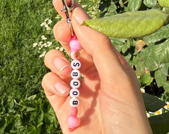 Puede incluir: Un llavero de cuentas con la palabra "BOOBS" escrita en letras blancas sobre cuentas rosas. También son visibles dos pulseras de cuentas, una con "GAY ASF" y la otra con "FRUITY" escrita en cuentas coloridas.