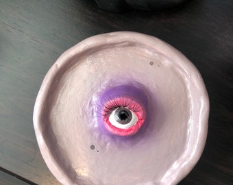 Puede incluir: Un cuenco para baratijas redondo hecho a mano, de color lavanda, con un ojo esculpido en el centro. El ojo tiene una pupila negra, labios rosas y largas pestañas moradas. Una pieza artística única.