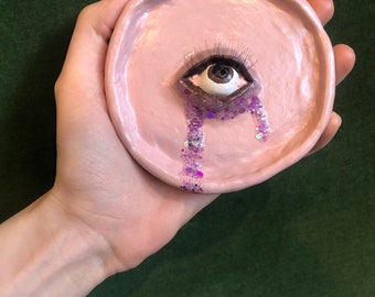 Puede incluir: Un platillo de cerámica rosa con un ojo detallado, largas pestañas y lágrimas de purpurina morada. El plato redondo se sostiene en una mano, mostrando un diseño artístico. El ojo tiene un iris marrón y es el elemento central de la pieza.