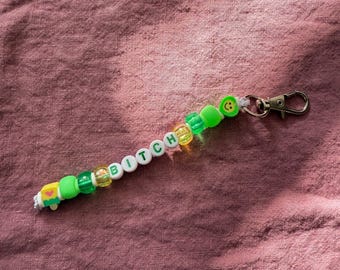 Puede incluir: Un llavero de cuentas con la palabra "BITCH" escrita con cuentas de letras blancas. El llavero presenta cuentas verdes, amarillas y transparentes, junto con una cuenta de carita sonriente y un cierre plateado. El fondo es una tela malva texturizada.
