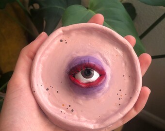 Puede incluir: Un plato de cerámica rosa hecho a mano con un ojo pintado, labios rojos, sombra de ojos morada y pestañas negras. El plato redondo tiene pequeñas motas oscuras y está sostenido en una mano.