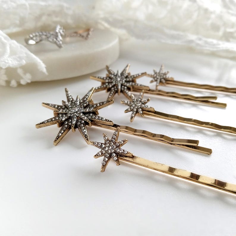 May include: A set of gold-toned hair pins featuring star-shaped ornaments encrusted with clear rhinestones. The pins have a wavy design and vary in size, with each star radiating multiple points. The metal finish is a polished metallic gold.