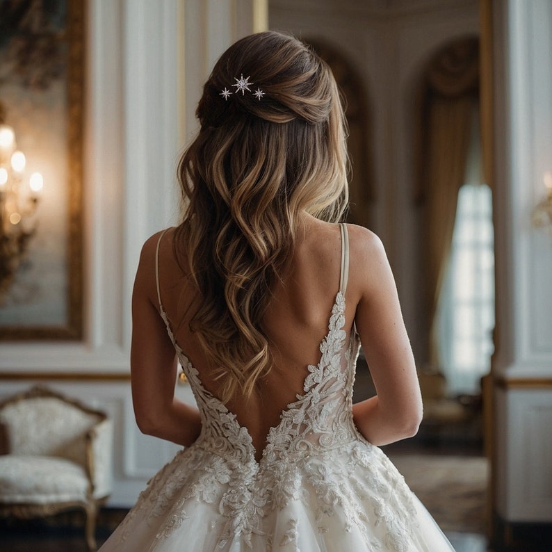 May include: A white sleeveless gown with a deep V-back, featuring intricate floral lace embroidery and a full skirt. The hair is styled in a half-up design, secured with a silver star-shaped hair accessory with small clear crystals.