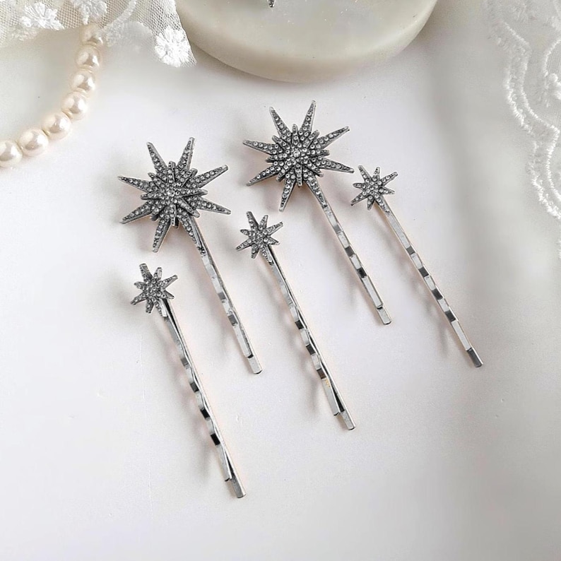 May include: A set of four silver-toned hair pins, each featuring a starburst-shaped ornament encrusted with small clear rhinestones. The pins vary in size, with two larger starburst designs and two smaller starburst designs attached to metal bobby pins.