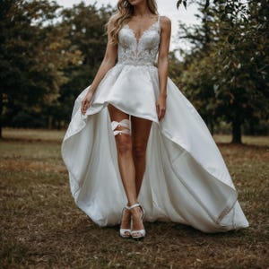 K&ouml;nnte beinhalten: Ein wei&szlig;es Hochzeitskleid mit einem hoch-tiefen Saum und einem Spitzenoberteil. Das Kleid hat einen Satinrock und eine zarte Spitzenstrumpfband am Bein.