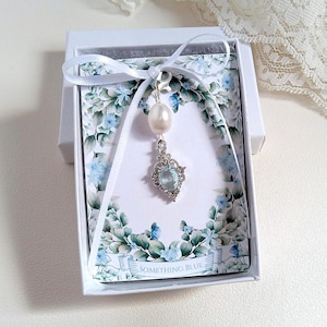 Puede incluir: Un dije plateado con una perla blanca y un cristal ovalado azul claro en un engaste decorativo, sujeto a una cinta blanca. Se presenta en una caja blanca sobre una tarjeta con borde floral y el texto SOMETHING BLUE.