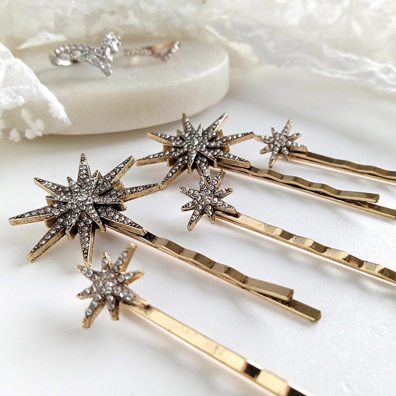 May include: A collection of gold-toned bobby pins featuring starburst-shaped ornaments encrusted with clear rhinestones. The hair accessories vary in size, with larger and smaller star designs attached to the ends of the metallic wavy pins.
