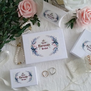 K&ouml;nnte beinhalten: Vier wei&szlig;e Boxen mit dem Text "Love Forever Bridal" darauf. Die Boxen sind mit floralen Designs verziert und auf einer wei&szlig;en Oberfl&auml;che angeordnet. Zwei goldene Ringe sind ebenfalls auf dem Bild zu sehen.