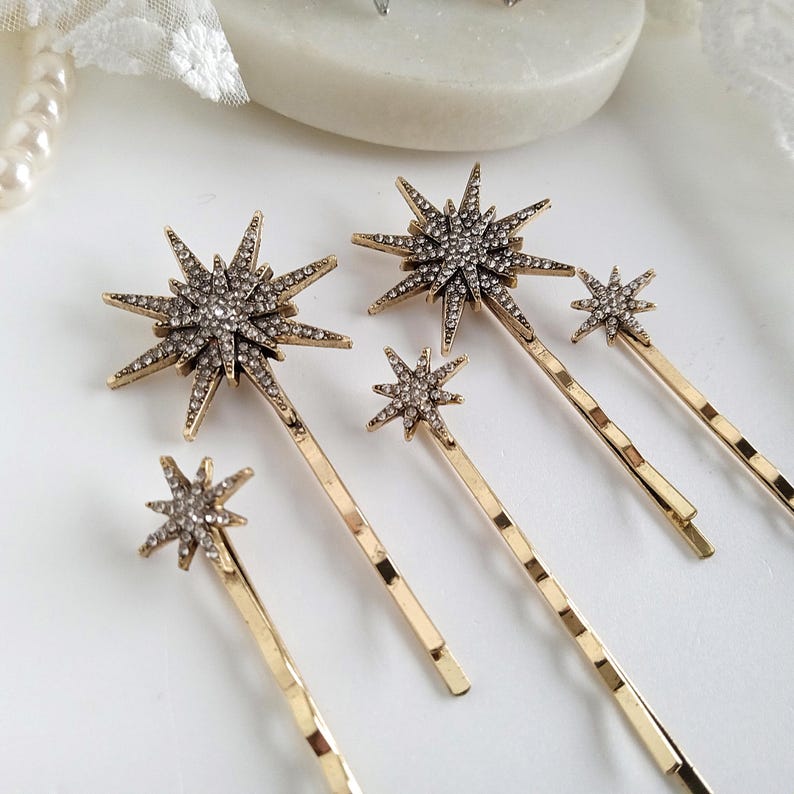 May include: A set of four gold-toned hair pins, each featuring a starburst-shaped ornament encrusted with small clear rhinestones. The starbursts vary in size, with two larger designs and two smaller designs, all attached to simple metal hair clips.