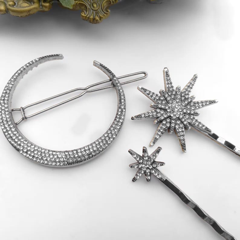 May include: A set of three silver-toned hair accessories featuring a large crescent moon shape and two star-shaped designs, all encrusted with small clear rhinestones. Each piece is attached to a thin, metallic hair pin for secure placement.