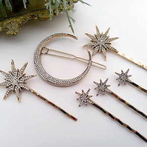 May include: A gold-toned hair accessory set featuring one large crescent moon hair clip encrusted with clear rhinestones and five star-shaped hair pins of varying sizes, each decorated with clear rhinestones on a gold-toned metal base.