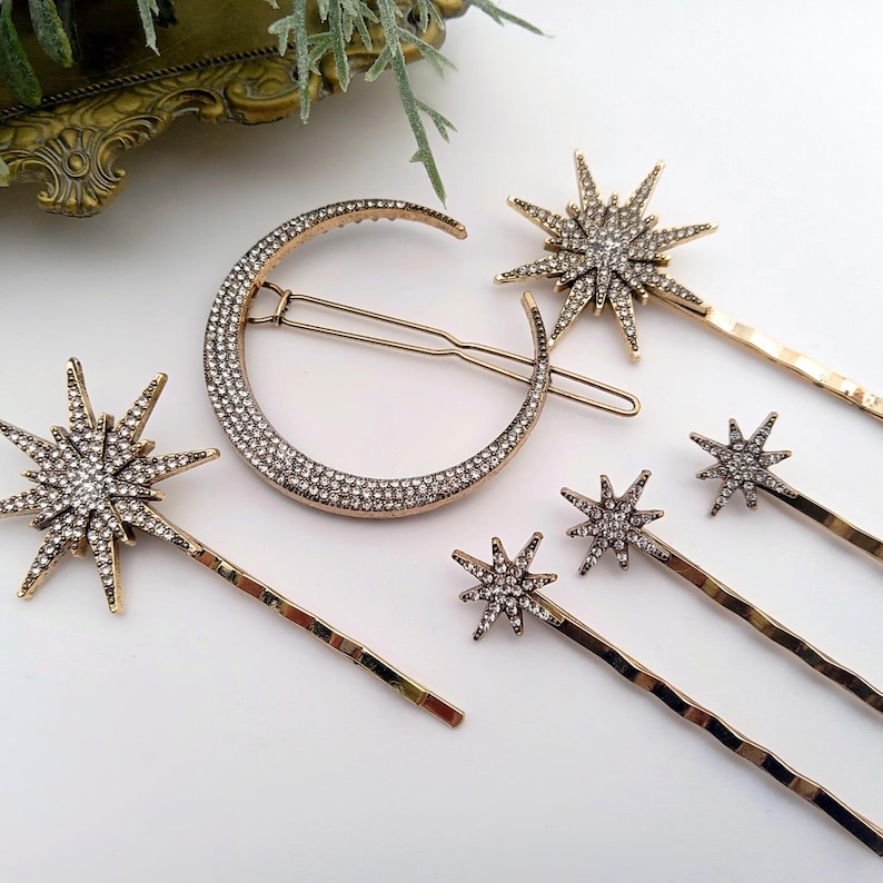 May include: A gold-toned hair accessory set featuring one large crescent moon hair clip encrusted with clear rhinestones and five star-shaped hair pins of varying sizes, each decorated with clear rhinestones on a gold-toned metal base.