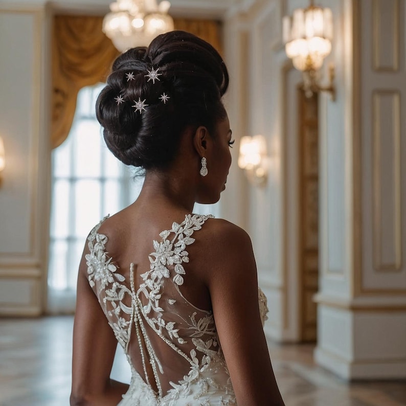May include: A sleeveless white gown featuring intricate floral lace embroidery on the bodice and a sheer back panel adorned with delicate pearl strands. The hair is styled in an elegant updo accented with small, sparkling star-shaped hair pins.