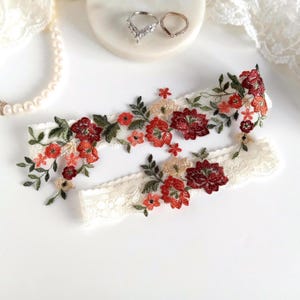 May include: Ivory lace garter set with embroidered red and peach floral design. The floral pattern includes green leaves and small flowers. Two rings are on a white surface, and a pearl necklace is in the background.
