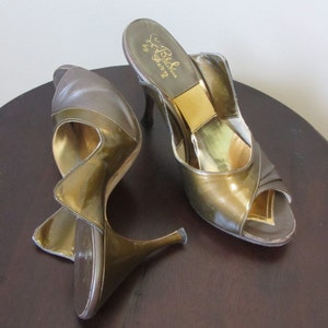 May include: A pair of vintage open-toe high heels in olive green and gold. The shoes have a unique, sculptural design with a curved heel and a metallic gold interior. The heels are displayed on a dark brown, round table.