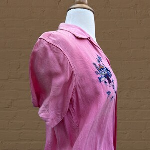 May include: A pink short-sleeved button-down shirt with an embroidered design on the front. The design features a blue bird, a yellow sun, and green plants.