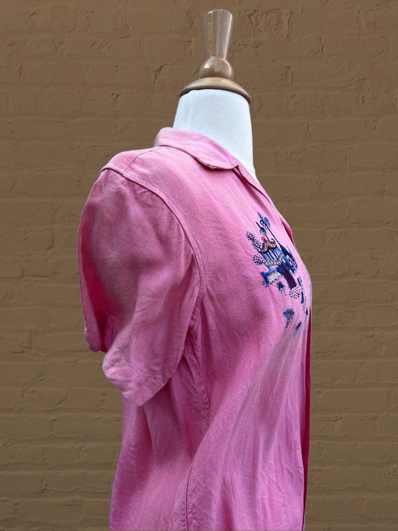 May include: A pink short-sleeved button-down shirt with an embroidered design on the front. The design features a blue bird, a yellow sun, and green plants.