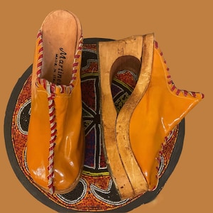 May include: A pair of orange leather clogs with red and white stitching. The clogs are on a wooden platform and have a label that reads "Martine" and "Made in Italy".