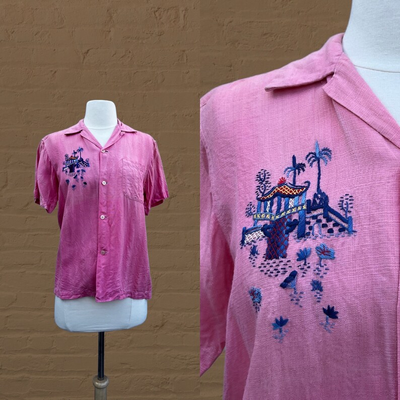 May include: A pink short-sleeved button-up shirt with blue and red embroidered details of a pagoda and palm trees.