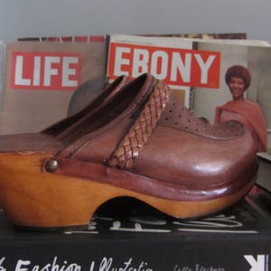 May include: Brown leather platform clogs with a woven strap detail. The clogs are sitting on top of a stack of magazines, including "LIFE" and "EBONY".