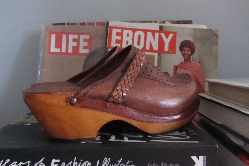 May include: Brown leather platform clogs with a woven strap detail. The clogs are sitting on top of a stack of magazines, including "LIFE" and "EBONY".