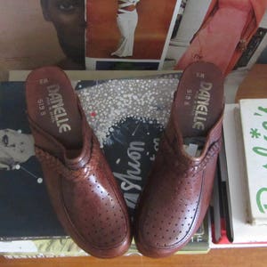 May include: A pair of brown leather clogs with a woven strap detail and perforated design. The clogs have a gold label that reads "DANELLE 6M 5198 B Made in Brazil".