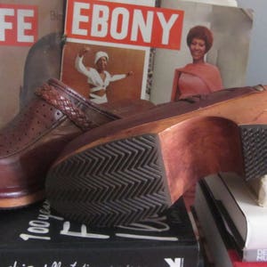 May include: A pair of brown leather clogs with a wooden platform sole. The clogs have a woven strap detail and are sitting on a stack of books. The books have titles like "Life", "Ebony", "Barbie's", and "The Black Californians".