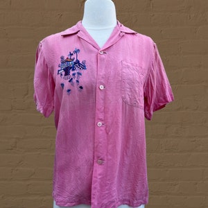May include: A pink short-sleeved button-down shirt with an embroidered scene of a tropical island with palm trees and a building. The shirt has a pocket on the left side.