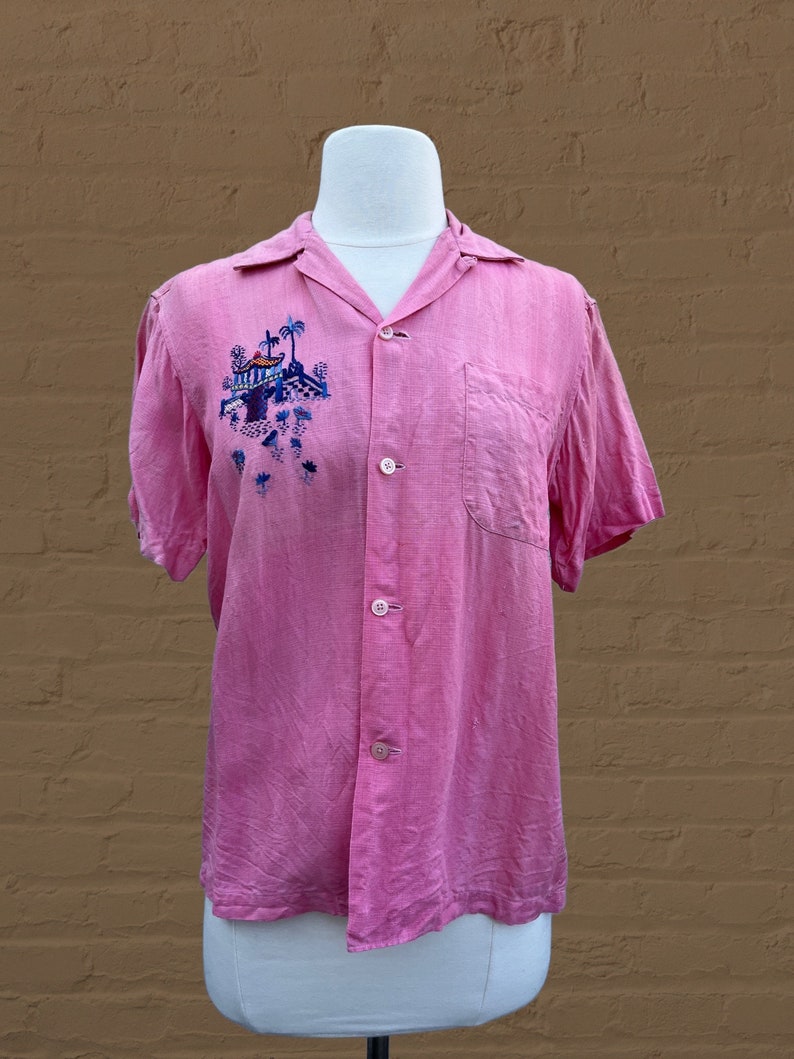 May include: A pink short-sleeved button-down shirt with an embroidered scene of a tropical island with palm trees and a building. The shirt has a pocket on the left side.