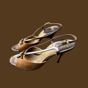 K&ouml;nnte beinhalten: Ein Paar braune Wildledersandalen mit Absatz, goldener Schnalle und goldenem Riemen. Die Sandalen haben eine offene Zehenpartie und einen Slingback-Riemen.