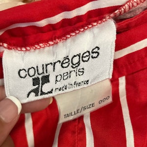 May include: White label with black text reading "Courr&egrave;ges Paris" and "Made in France". Below the label is a white tag with black text reading "Taille/Size 000". The label is attached to a red and white striped fabric.