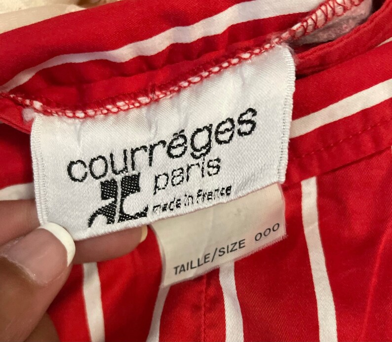 May include: White label with black text reading "Courr&egrave;ges Paris" and "Made in France". Below the label is a white tag with black text reading "Taille/Size 000". The label is attached to a red and white striped fabric.