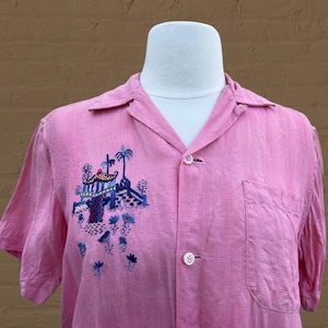 May include: A pink short-sleeved button-up shirt with an embroidered design of a pagoda and palm trees in blue, green, and yellow.