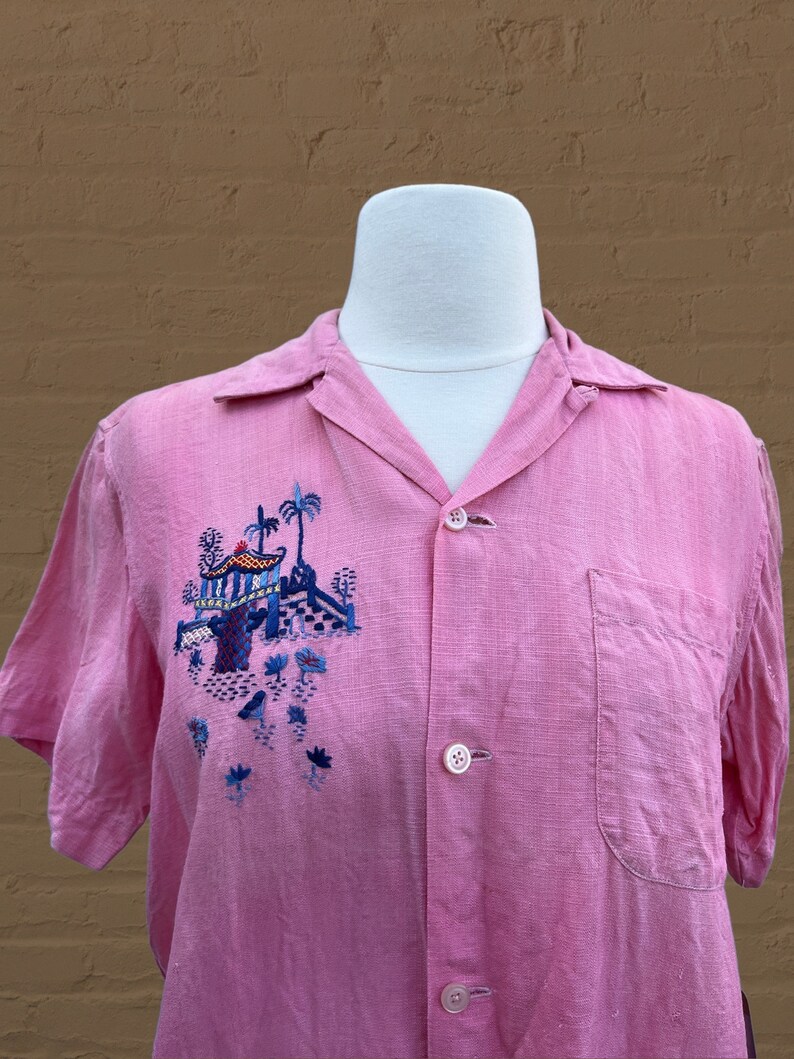 May include: A pink short-sleeved button-up shirt with an embroidered design of a pagoda and palm trees in blue, green, and yellow.