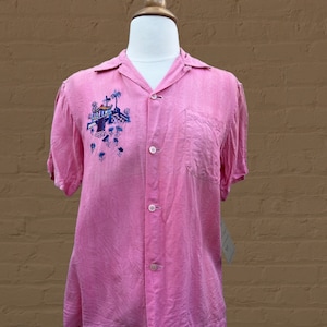 May include: A pink short-sleeved button-down shirt with an embroidered design of a building with palm trees on the left side of the chest.