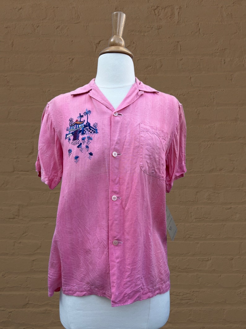 May include: A pink short-sleeved button-down shirt with an embroidered design of a building with palm trees on the left side of the chest.