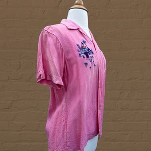 May include: A pink short-sleeved button-up shirt with an embroidered design of a tropical scene with palm trees and a building. The shirt is on a white mannequin.