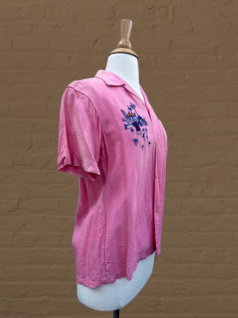 May include: A pink short-sleeved button-up shirt with an embroidered design of a tropical scene with palm trees and a building. The shirt is on a white mannequin.