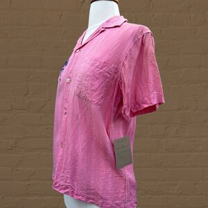May include: A pink short-sleeved button-up shirt with a small embroidered design on the left side of the chest. The shirt is on a white mannequin.