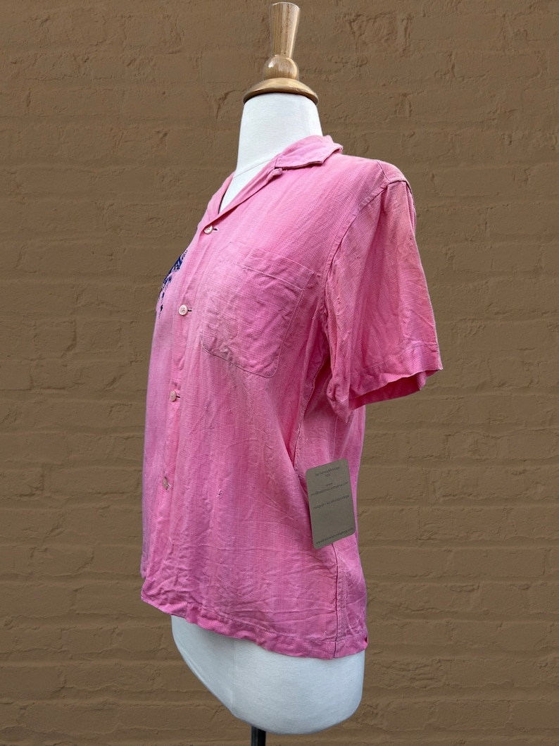 May include: A pink short-sleeved button-up shirt with a small embroidered design on the left side of the chest. The shirt is on a white mannequin.