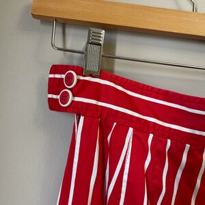May include: Red and white striped skirt with a button closure at the waist. The skirt is pleated and has a wide waistband.