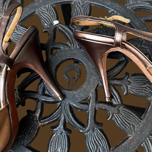 K&ouml;nnte beinhalten: Ein Paar braune Ledersandalen mit Absatz und Kn&ouml;chelriemen, die auf einer schwarzen Metalloberfl&auml;che mit einem dekorativen Blumenmuster liegen.