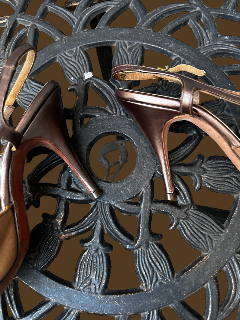 K&ouml;nnte beinhalten: Ein Paar braune Ledersandalen mit Absatz und Kn&ouml;chelriemen, die auf einer schwarzen Metalloberfl&auml;che mit einem dekorativen Blumenmuster liegen.