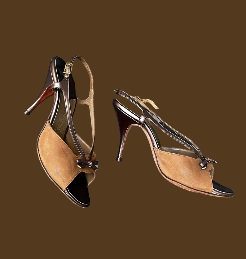 K&ouml;nnte beinhalten: Ein Paar braune Wildledersandalen mit Absatz, goldener Schnalle und schwarzem Lederriemen. Die Sandalen haben eine offene Zehenpartie und ein Slingback-Design.