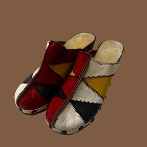 May include: A pair of colorful clogs with a patchwork design. The clogs feature red, black, white, and yellow leather panels stitched together. The soles are made of wood and have a slight heel.